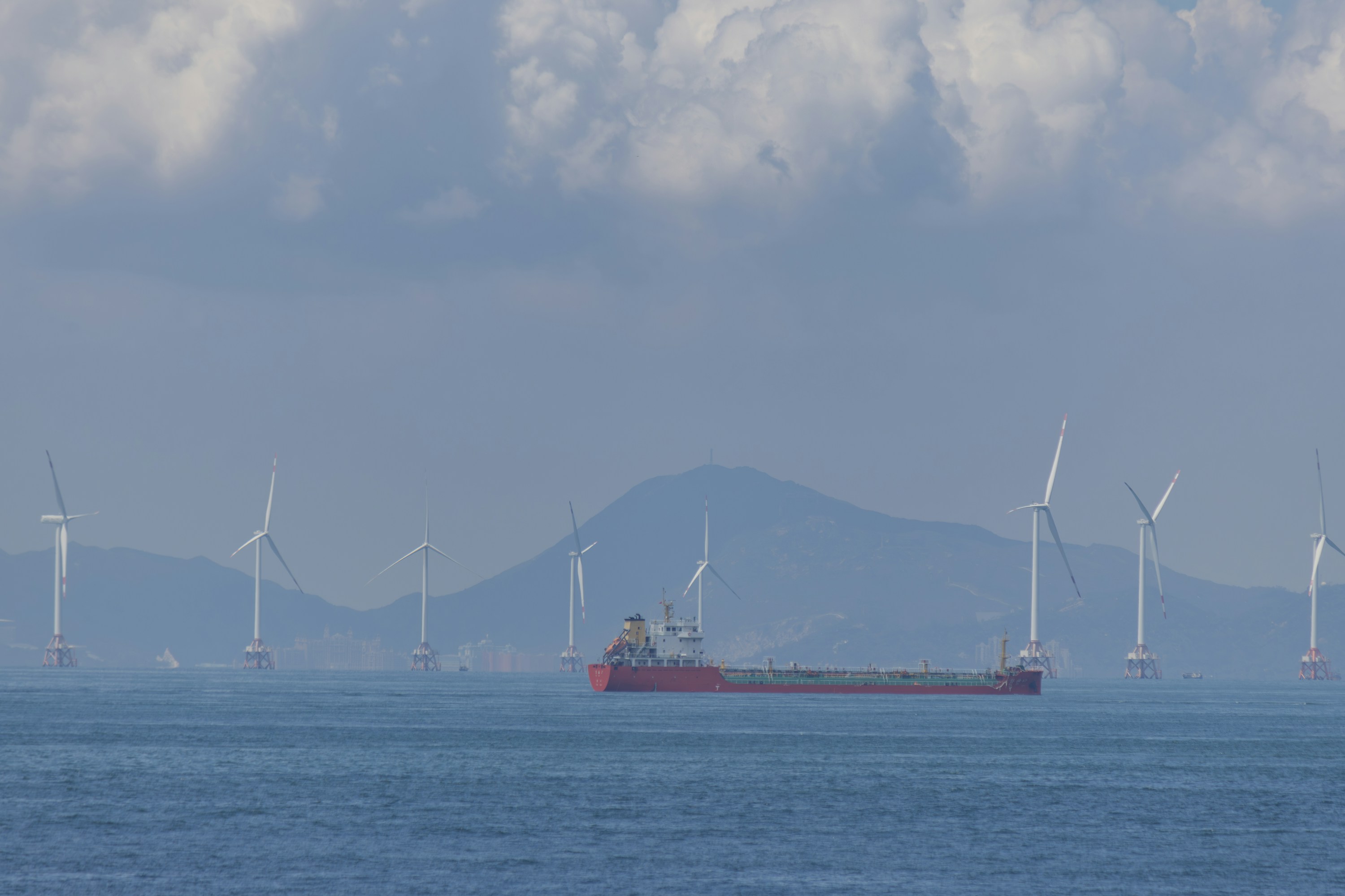A red cargo vessel crossing calm water in front of offshore wind turbines and a hazy mountain coast